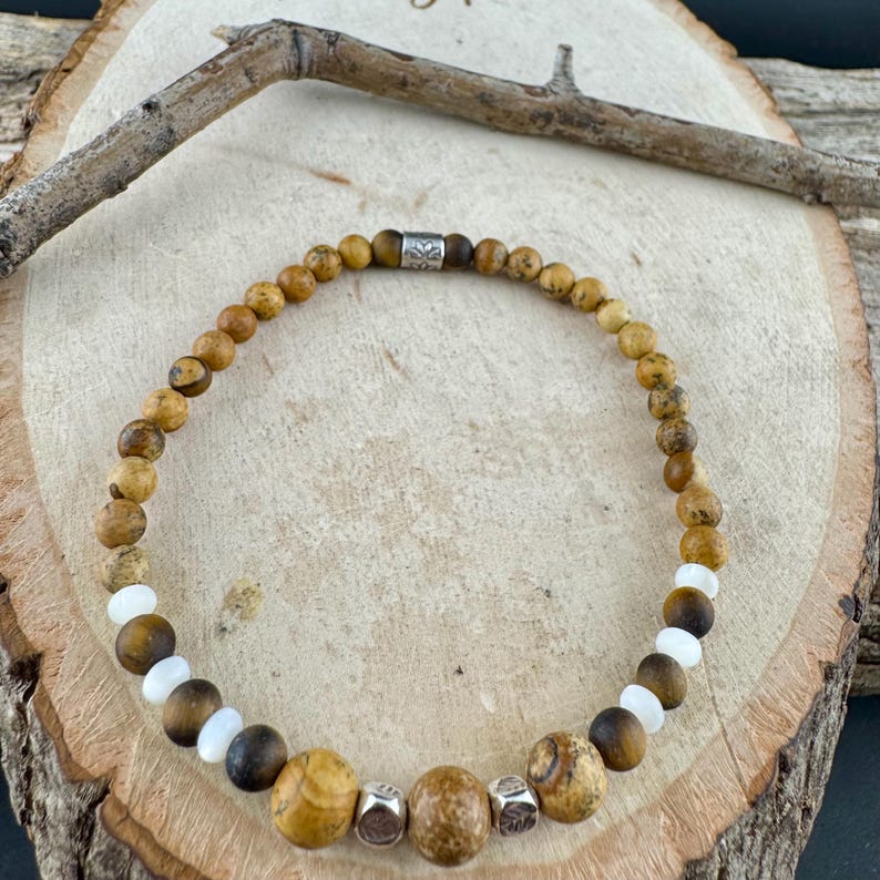 May include: A beaded bracelet featuring a mix of natural stone beads in shades of brown, tan, and white. The bracelet includes a few silver-colored metal beads. The bracelet is displayed on a wooden surface.