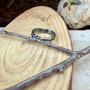 May include: A silver-toned metal ring featuring a textured, wavy band design with dark oxidised detailing in the grooves. The ring is positioned resting on a thin, brown wooden twig, which is placed across a light-coloured, oval-shaped wood slice.