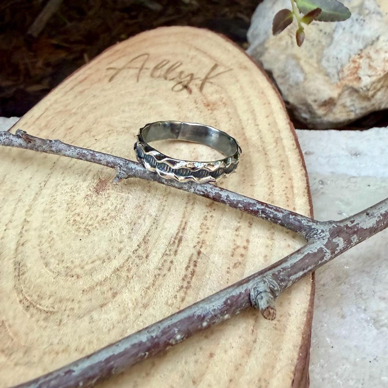 May include: A silver-toned metal ring featuring a textured, wavy band design with dark oxidised detailing in the grooves. The ring is positioned resting on a thin, brown wooden twig, which is placed across a light-coloured, oval-shaped wood slice.