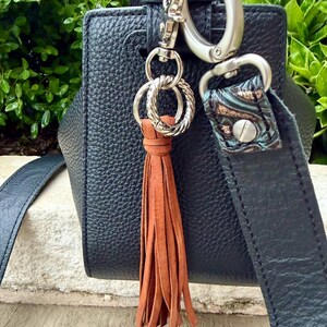 May include: Close-up of a black leather handbag with a brown leather tassel. The bag has silver-tone hardware, including a clasp and decorative rings. The strap features a patterned accent.