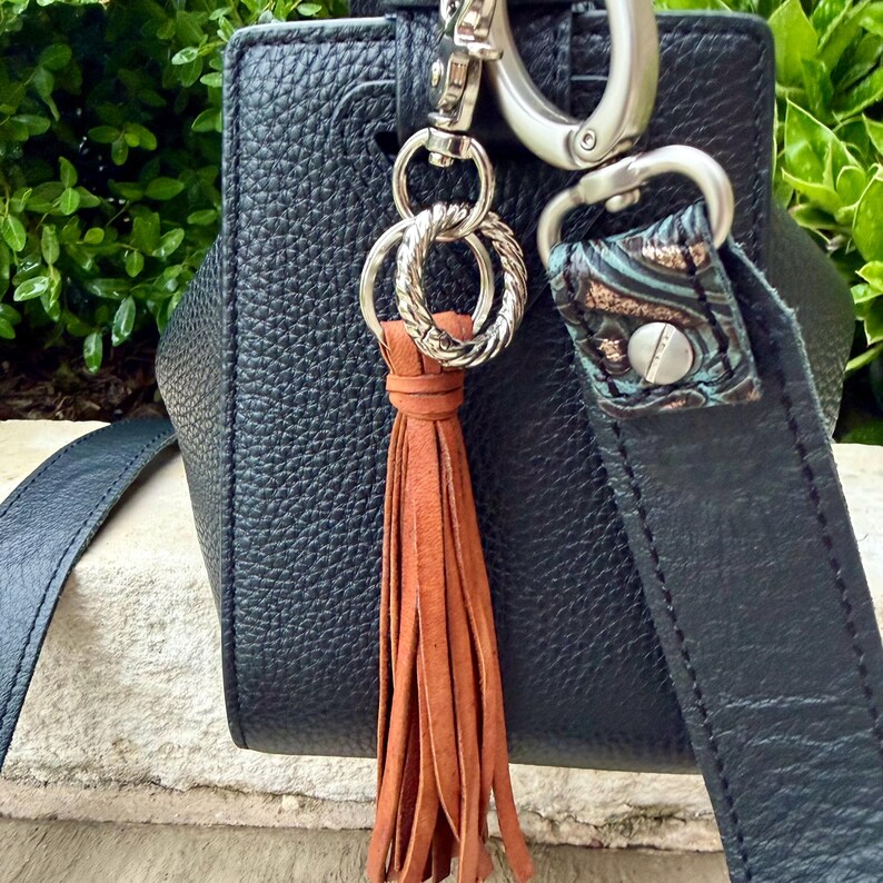 May include: Close-up of a black leather handbag with a brown leather tassel. The bag has silver-tone hardware, including a clasp and decorative rings. The strap features a patterned accent.