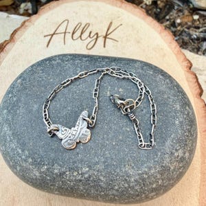Op de afbeelding: Een zilverkleurige ketting met een decoratieve hanger. De hanger heeft een sierlijk ontwerp. De ketting wordt gepresenteerd op een grijze steen, met de naam "Allyk" geschreven op een houten oppervlak op de achtergrond.