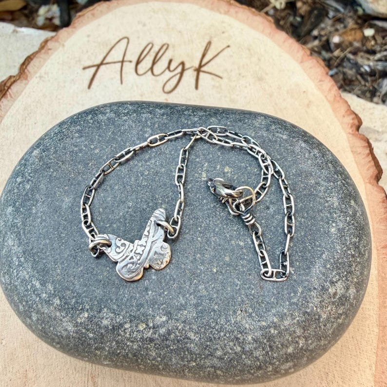 Op de afbeelding: Een zilverkleurige ketting met een decoratieve hanger. De hanger heeft een sierlijk ontwerp. De ketting wordt gepresenteerd op een grijze steen, met de naam "Allyk" geschreven op een houten oppervlak op de achtergrond.