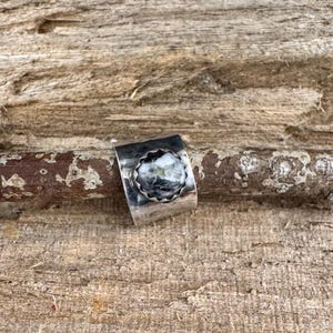 May include: A silver ring with a round, textured, black and white stone set in a scalloped bezel. The ring band is wide and has a hammered texture. The ring is displayed on a piece of wood.