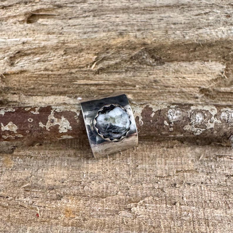May include: A silver ring with a round, textured, black and white stone set in a scalloped bezel. The ring band is wide and has a hammered texture. The ring is displayed on a piece of wood.