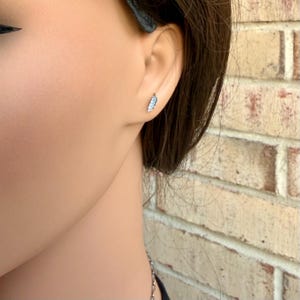 May include: A close-up of a silver feather earring on a person's ear. The earring is small and detailed, with a delicate feather design. The background is a brick wall.