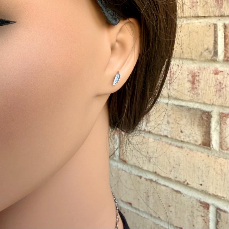 May include: A close-up of a silver feather earring on a person's ear. The earring is small and detailed, with a delicate feather design. The background is a brick wall.