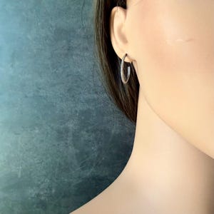 May include: A close-up of a silver hoop earring. The earring is a simple, circular design with a textured surface. The earring is worn on the ear of a person with long, dark hair. The background is a muted teal color.