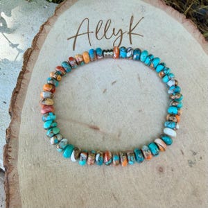 May include: A circular beaded bracelet featuring small, flat, round stones in shades of turquoise, orange, white, and brown with visible matrix patterns. The bracelet includes a single metallic silver-toned spacer bead and is displayed on a light wood slice with the text Ally K written in dark brown script.