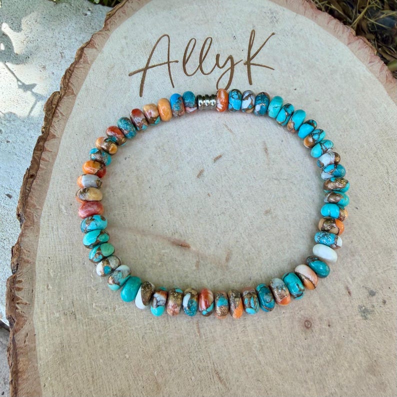 May include: A circular beaded bracelet featuring small, flat, round stones in shades of turquoise, orange, white, and brown with visible matrix patterns. The bracelet includes a single metallic silver-toned spacer bead and is displayed on a light wood slice with the text Ally K written in dark brown script.