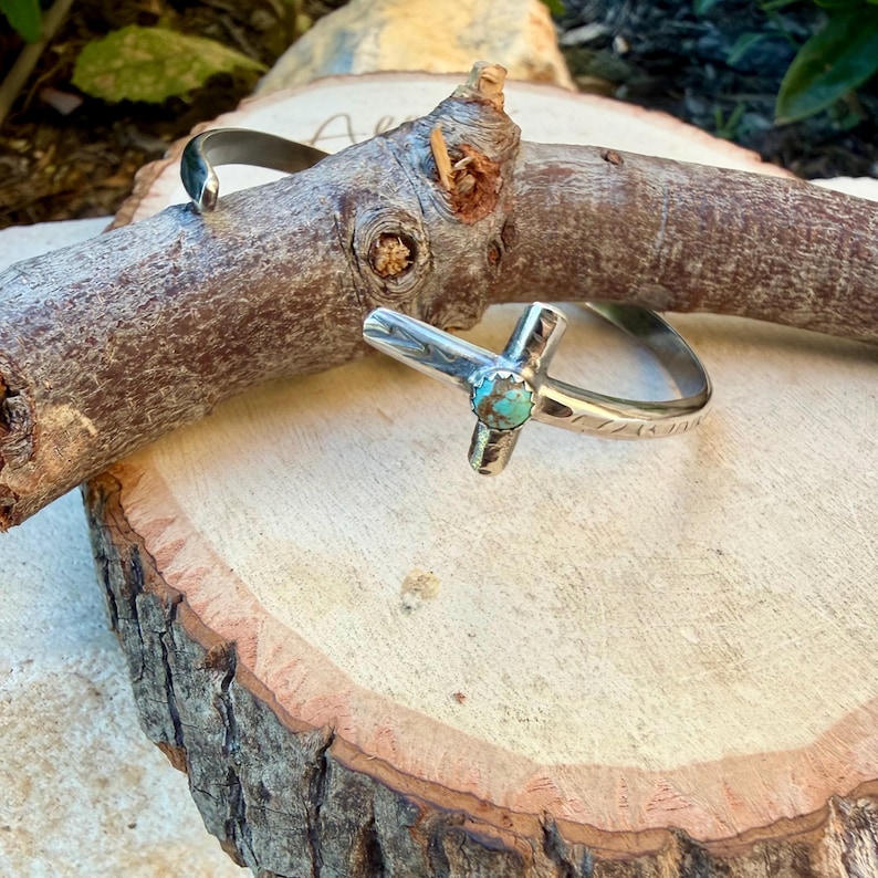 May include: Two silver rings. One is a simple band, the other features a cross design with a small turquoise stone at the center. The rings are displayed on a wooden surface, showcasing their handcrafted details.