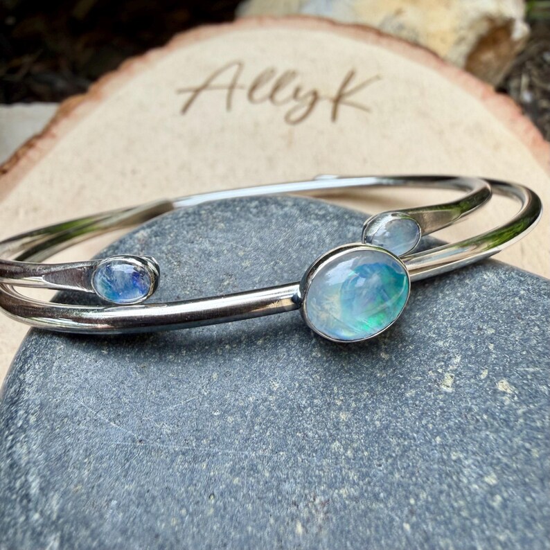 K&ouml;nnte beinhalten: Ein silberfarbenes Armband mit drei ovalen, irisierenden Edelsteinen. Das Armband liegt auf einem grauen Stein, mit einem Holzuntergrund, auf dem der Name "Allyk" eingraviert ist.
