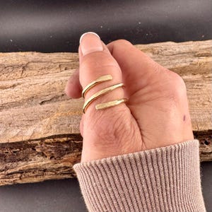 May include: A gold-colored, spiral ring with a hammered texture. The ring wraps around the finger with three distinct bands. The ring is shown on a hand with a neutral-toned sweater sleeve in the background.