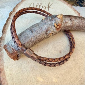 Puede incluir: Una pulsera de cuero trenzado marr&oacute;n de doble hebra con un cierre magn&eacute;tico cil&iacute;ndrico de tono cobre met&aacute;lico. El cord&oacute;n de cuero muestra tonos variados de marr&oacute;n claro y oscuro, creando una apariencia texturizada en toda la banda circular.