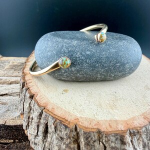 May include: A gold-colored open cuff bracelet with turquoise-colored stone accents. The bracelet is resting on a gray stone, which is placed on a wooden surface. The bracelet's design features a curved shape with a stone at each end.