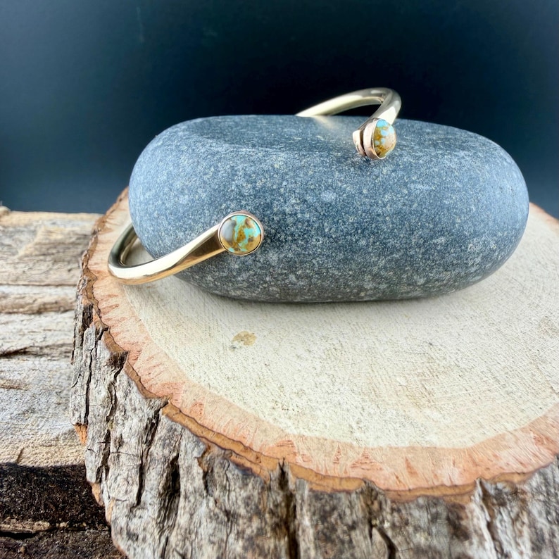 May include: A gold-colored open cuff bracelet with turquoise-colored stone accents. The bracelet is resting on a gray stone, which is placed on a wooden surface. The bracelet's design features a curved shape with a stone at each end.