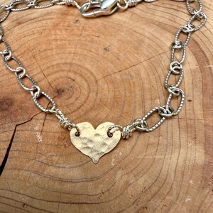 May include: A bracelet featuring a central gold-toned heart charm with a hammered, textured surface. The heart is connected on both sides to a silver-toned chain composed of oval-shaped links with a subtle etched pattern and a lobster clasp.