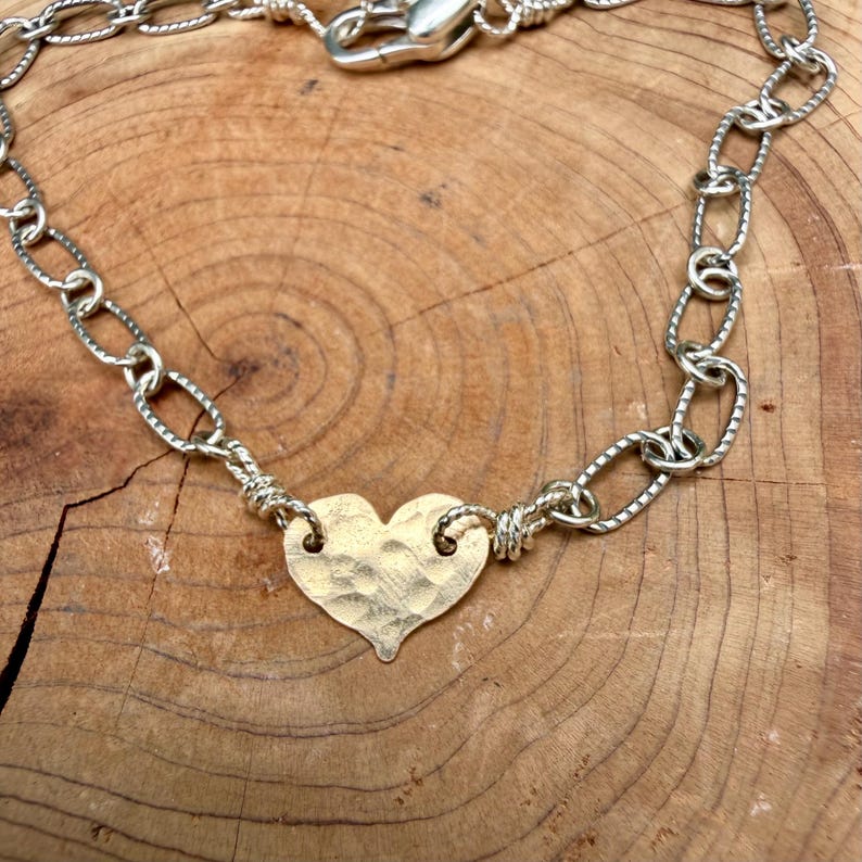 May include: A bracelet featuring a central gold-toned heart charm with a hammered, textured surface. The heart is connected on both sides to a silver-toned chain composed of oval-shaped links with a subtle etched pattern and a lobster clasp.