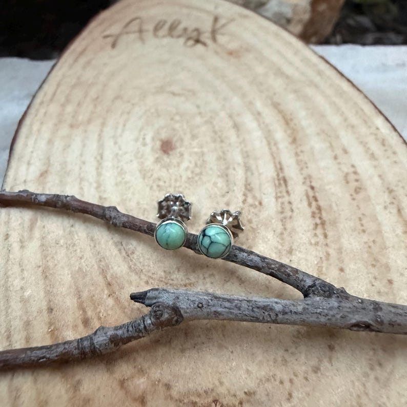 Pu&ograve; includere: Un paio di piccoli orecchini a lobo rotondi con una pietra verde chiaro marmorizzata incastonata in metallo argentato. Gli orecchini sono esposti su una piccola fetta di legno naturale con una firma scritta a mano nell'angolo in alto a sinistra.
