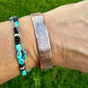 Puede incluir: Una pulsera de brazalete de cobre con un intrincado patr&oacute;n arremolinado se usa junto a una pulsera de cuentas turquesa y negras. El brazalete es de un color marr&oacute;n rojizo c&aacute;lido, y la pulsera de cuentas tiene detalles plateados.