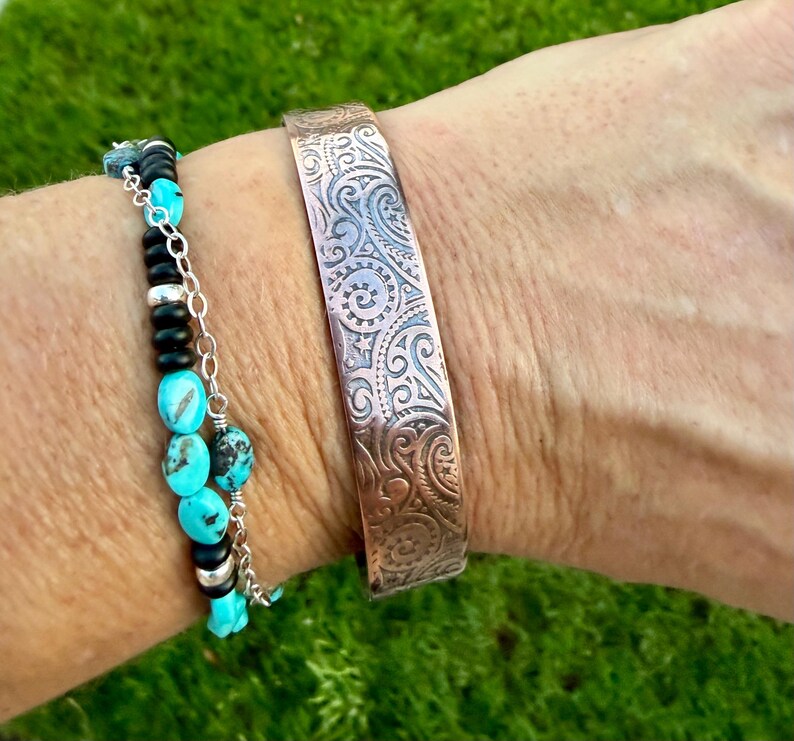 Puede incluir: Una pulsera de brazalete de cobre con un intrincado patr&oacute;n arremolinado se usa junto a una pulsera de cuentas turquesa y negras. El brazalete es de un color marr&oacute;n rojizo c&aacute;lido, y la pulsera de cuentas tiene detalles plateados.