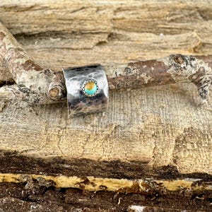 K&ouml;nnte beinhalten: Ein silberner Ring mit geh&auml;mmertem Muster und einem t&uuml;rkis- und goldfarbenen Stein. Der Ring ist vor einem nat&uuml;rlichen Holzgrund platziert und zeigt sein rustikales und handwerkliches Design. Das Band des Rings ist breit und umschlie&szlig;t den Finger.
