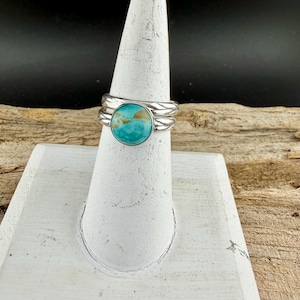 May include: A silver ring with a large, round turquoise stone. The ring has a textured band and is displayed on a white stand. The turquoise stone has a mix of blue and brown hues.