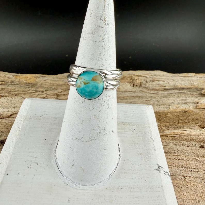 May include: A silver ring with a large, round turquoise stone. The ring has a textured band and is displayed on a white stand. The turquoise stone has a mix of blue and brown hues.