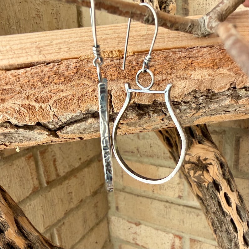 May include: A pair of silver earrings. One earring is a long, rectangular shape with etched details. The other earring is a curved, open shape resembling a stirrup. Both earrings hang from a simple hook.