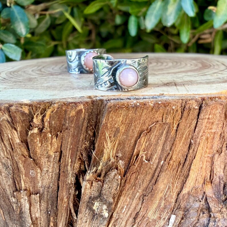 May include: Two silver-toned metal rings featuring a textured, hammered surface and a single, smooth, round, pale pink cabochon stone set in the centre of each band. The rings are positioned one behind the other on a wooden surface.