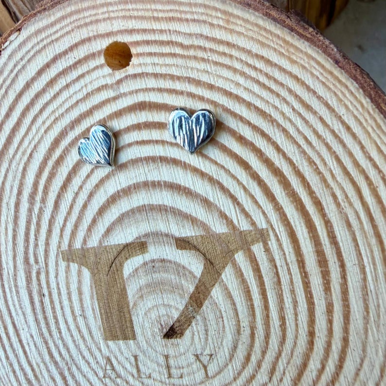 May include: A pair of small, silver-toned heart-shaped stud earrings with a textured, vertically lined surface. The earrings are displayed on a circular, light-colored wooden slice featuring a prominent wood grain pattern and the engraved text T7 ALLY.