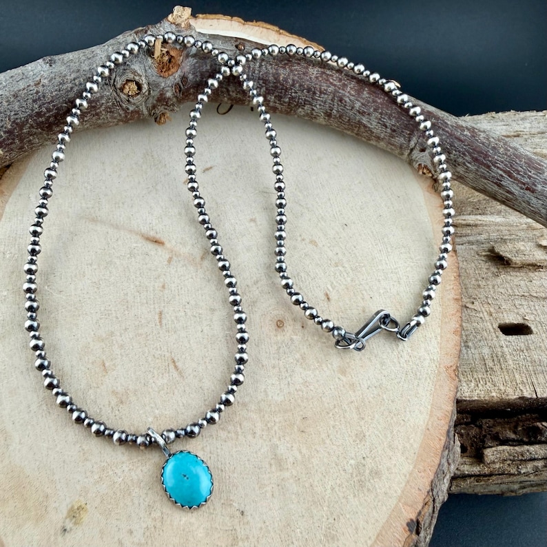 May include: A silver beaded necklace with a turquoise pendant. The pendant is an oval shape with a silver border. The necklace is displayed on a wooden surface with a dark background.