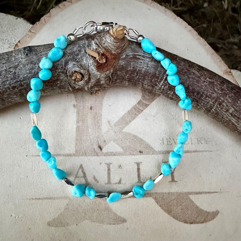 May include: A bracelet featuring small, irregular, bright blue turquoise-colored stone beads interspersed with thin, metallic silver-toned cylindrical spacers. The jewelry piece is secured with a silver-toned lobster claw clasp.