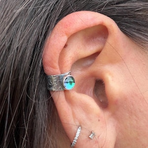 May include: A silver ear cuff with an etched design and a round, iridescent gemstone. The cuff is worn on the upper ear, showcasing the detailed metalwork and the colorful, reflective stone. Additional earrings are visible.
