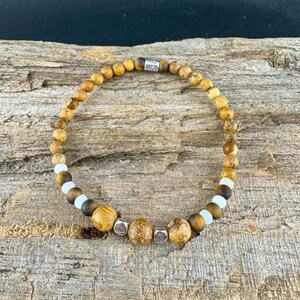 May include: A beaded bracelet featuring a mix of earth-toned beads, including speckled brown, tan, and white beads. The bracelet has a silver accent bead. The bracelet is displayed on a rustic wooden surface.