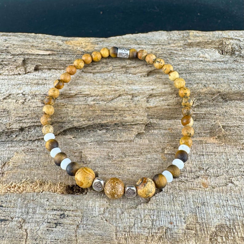 May include: A beaded bracelet featuring a mix of earth-toned beads, including speckled brown, tan, and white beads. The bracelet has a silver accent bead. The bracelet is displayed on a rustic wooden surface.