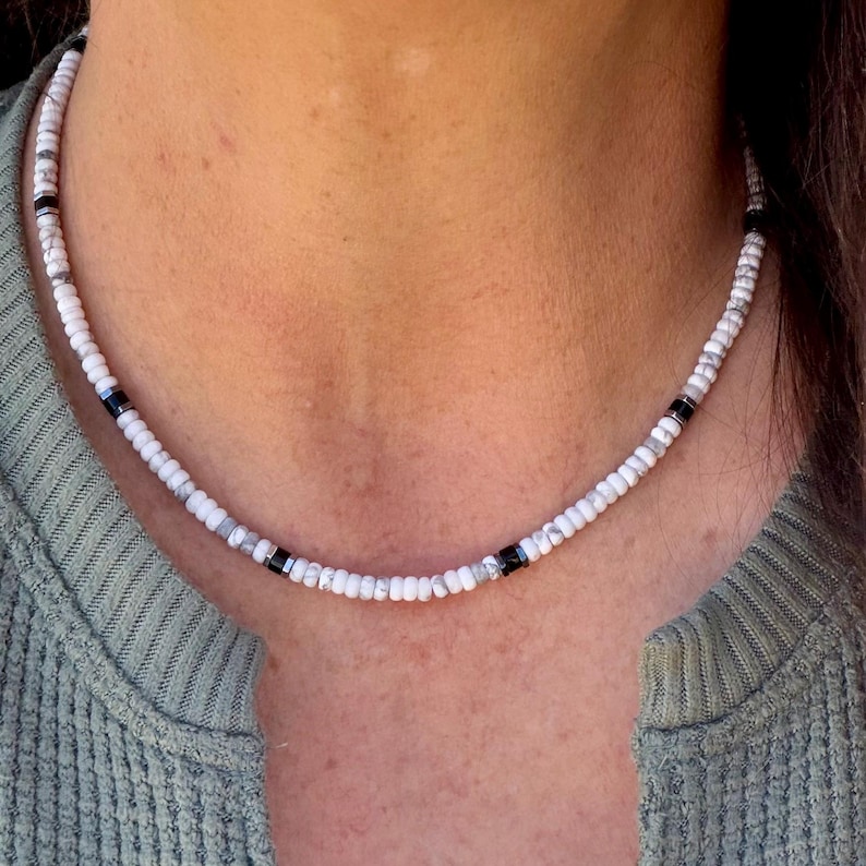 May include: White beaded necklace with black and gray accents. The necklace is composed of small, round, white beads interspersed with black and gray beads. The necklace is worn around the neck.