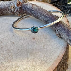 May include: A silver bangle bracelet with a round, turquoise-colored stone set in the center. The bracelet is resting on a wooden surface with a tree branch. The turquoise stone has a mottled appearance.