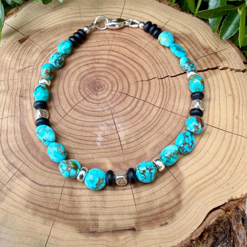May include: A turquoise beaded bracelet with silver and black accents. The bracelet features oval-shaped turquoise beads interspersed with silver-colored beads and black beads. The bracelet is displayed on a wooden surface.
