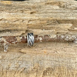 May include: A silver ring with an intricate, interwoven design. The ring is set against a rustic, wooden background, highlighting its detailed craftsmanship. The ring's design features a series of intertwined patterns, creating a textured appearance.