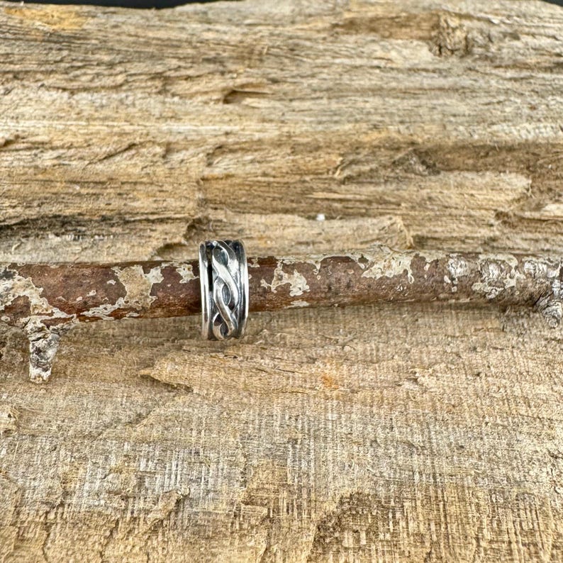 May include: A silver ring with an intricate, interwoven design. The ring is set against a rustic, wooden background, highlighting its detailed craftsmanship. The ring's design features a series of intertwined patterns, creating a textured appearance.