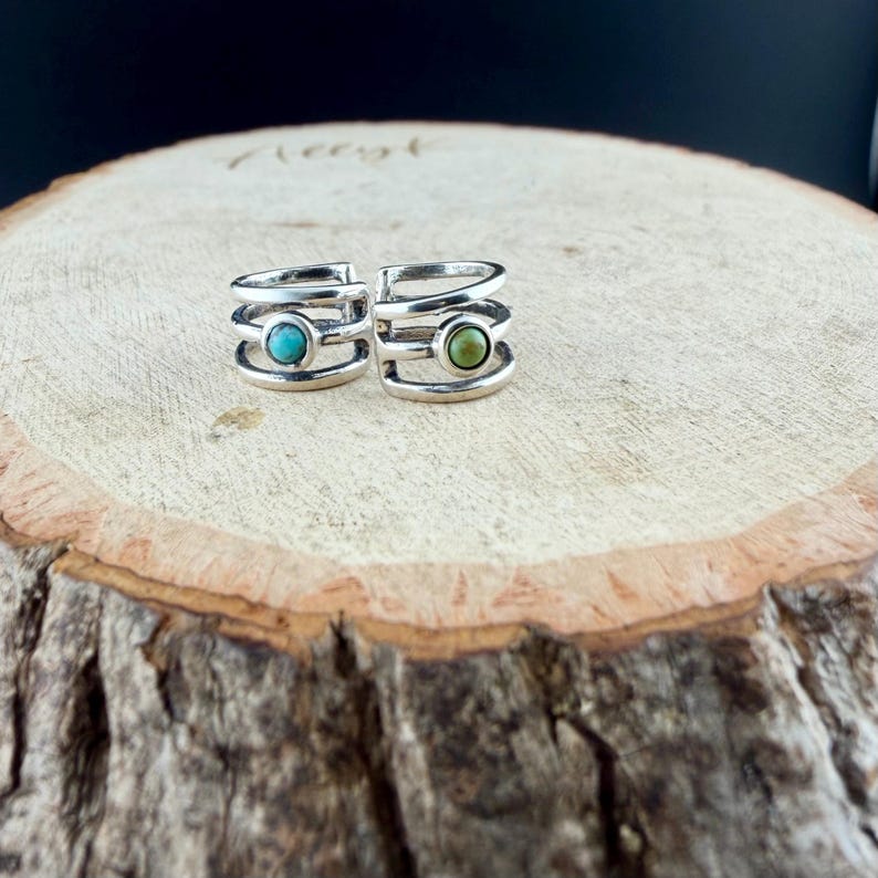 May include: Two silver-colored rings with a layered design. One ring features a turquoise-colored stone, and the other has a green stone. The rings are displayed on a wooden surface with a natural wood grain.