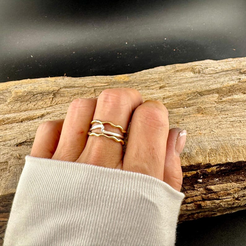 Puede incluir: Un anillo de dos tonos con un dise&ntilde;o ondulado. El anillo presenta una banda dorada en la parte superior y una banda plateada en la parte inferior. El anillo se lleva en un dedo, con una manga blanca en el fondo.
