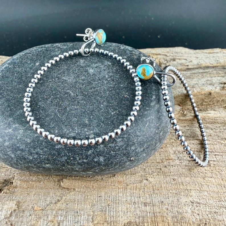 Puede incluir: Un par de pendientes de aro plateados y una pulsera a juego. Los pendientes y la pulsera est&aacute;n adornados con peque&ntilde;as cuentas redondas plateadas. Cada pendiente presenta una piedra de color turquesa y dorado. La joyer&iacute;a se exhibe sobre una superficie de piedra gris.