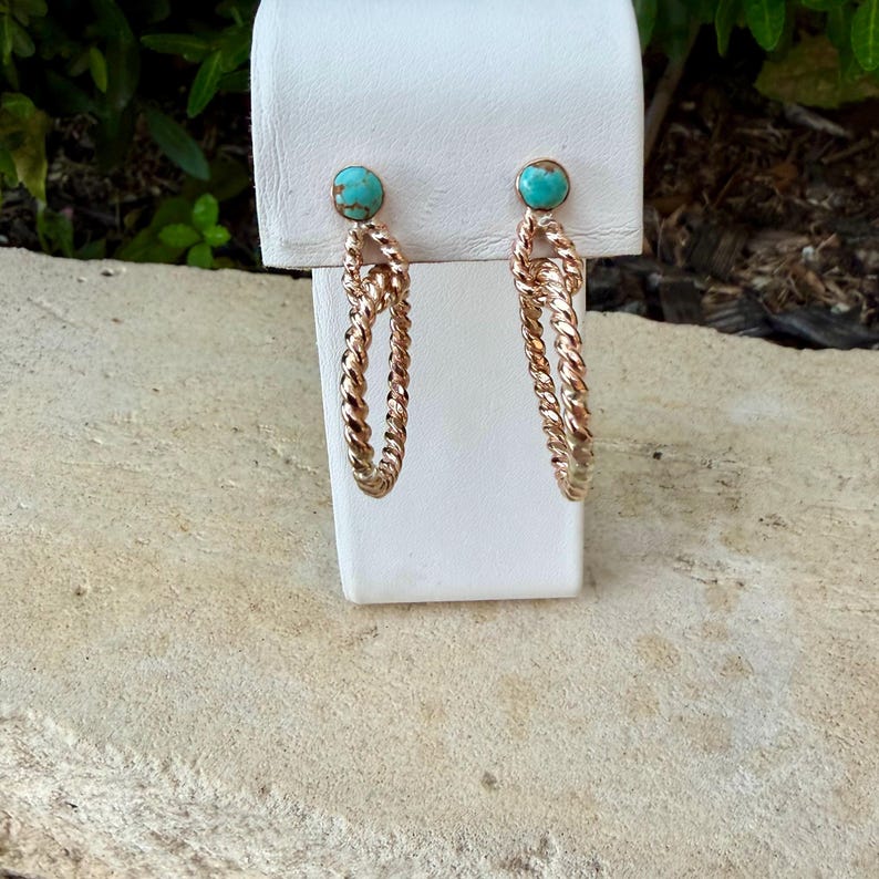 May include: A pair of gold-colored hoop earrings with a twisted rope design. Each earring features a small, round turquoise stone at the top. The earrings are displayed on a white stand against a natural background.