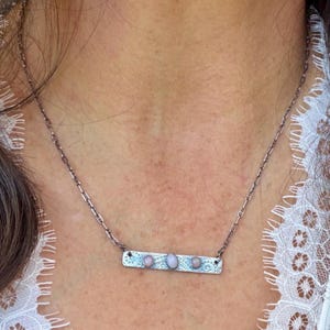 May include: A silver-toned metal bar necklace featuring three small, oval-shaped, light pink cabochon stones set horizontally. The rectangular bar has an etched, intricate pattern and is suspended from a delicate, thin metal chain.