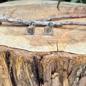 May include: A pair of silver-toned stud earrings shaped like hearts. Each earring features a textured, metallic surface with subtle, irregular lines and a polished finish. The earrings are positioned upright, showcasing their heart-shaped design and the small post backing visible behind the main face of the jewelry.