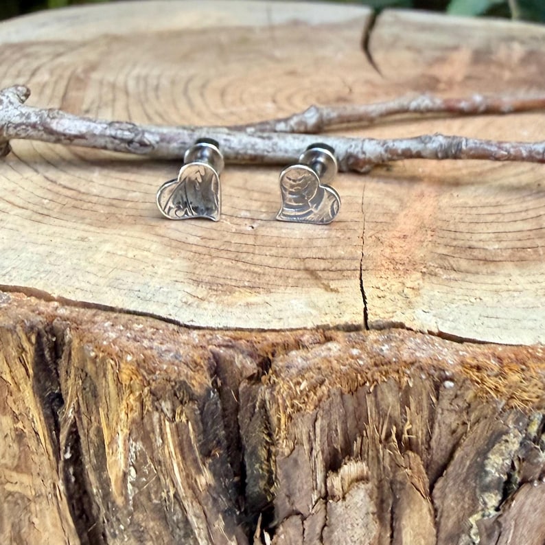 May include: A pair of silver-toned stud earrings shaped like hearts. Each earring features a textured, metallic surface with subtle, irregular lines and a polished finish. The earrings are positioned upright, showcasing their heart-shaped design and the small post backing visible behind the main face of the jewelry.