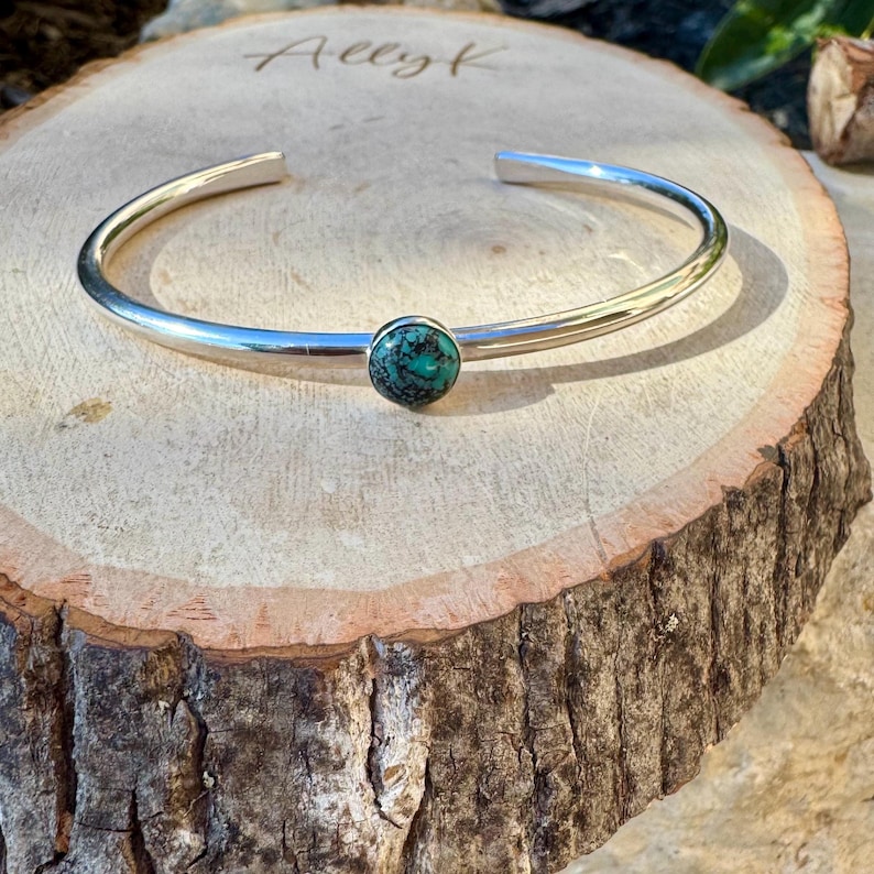 May include: A silver cuff bracelet with a turquoise stone set in the center. The bracelet is displayed on a wooden surface. The word "Allyk" is engraved on the wood.