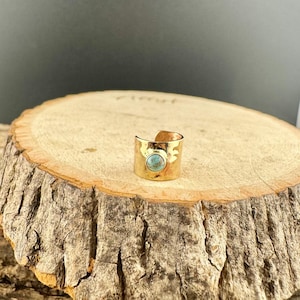 May include: A gold-colored ring with a turquoise-colored stone. The ring has a wide band with a textured surface and decorative cutouts. The turquoise stone is round and set in the center of the ring.